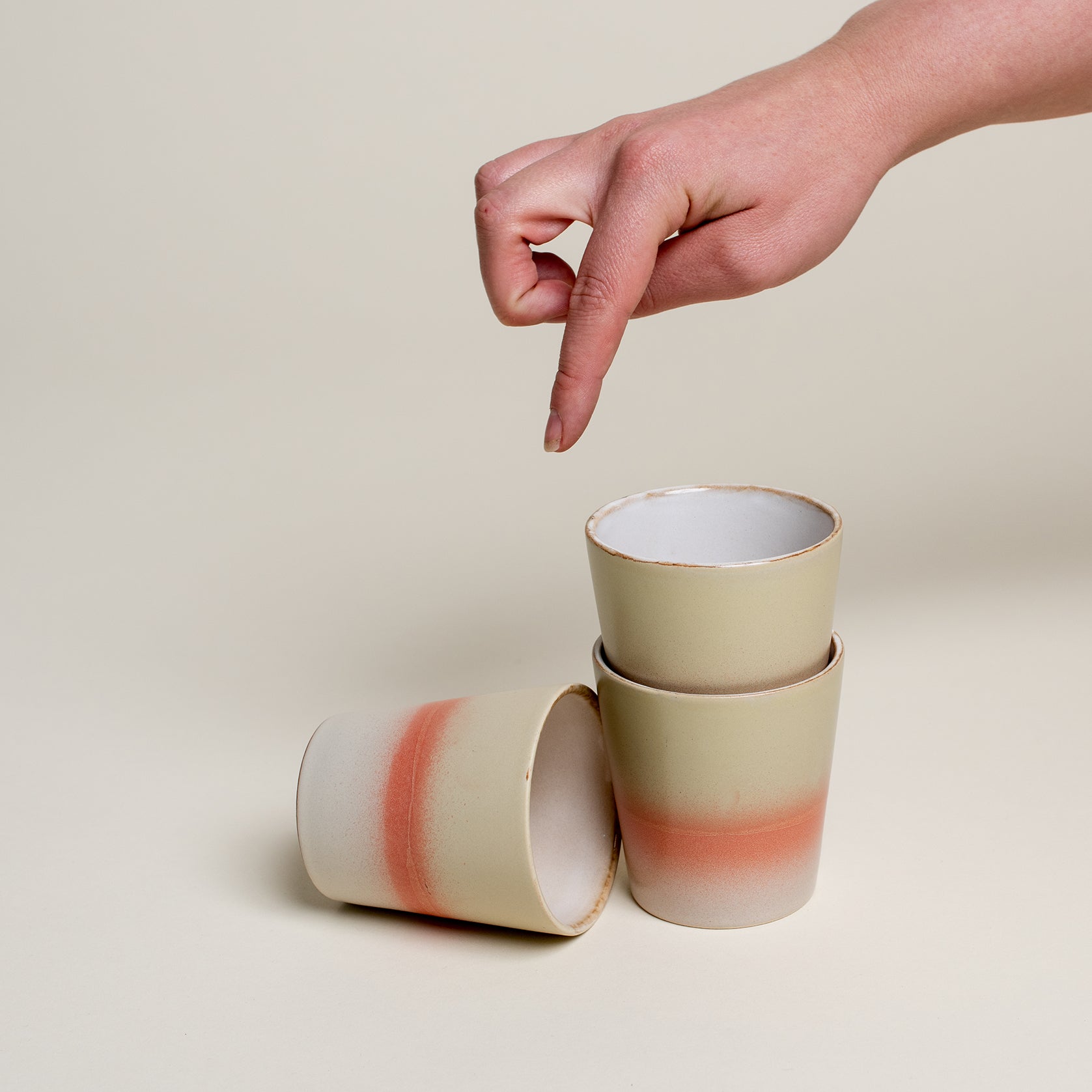  Tee Becher sind unten hell grau, in der Mitte orange und oben pastelle grün gefärbt. Eine Person zeigt von oben auf die Tassen.