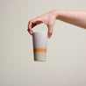 Eine Person hält eine Tasse in die Luft. Die Tasse im 70er retro Stil ist unten hell grün, in der mitte orange und oben hell grau gefärbt.