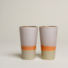 Zwei Tassen stehen in einem weißen Hintergrund nebeneinander. Die Becher im 70er retro Stil ist unten hell grün, in der mitte orange und oben hell grau gefärbt.