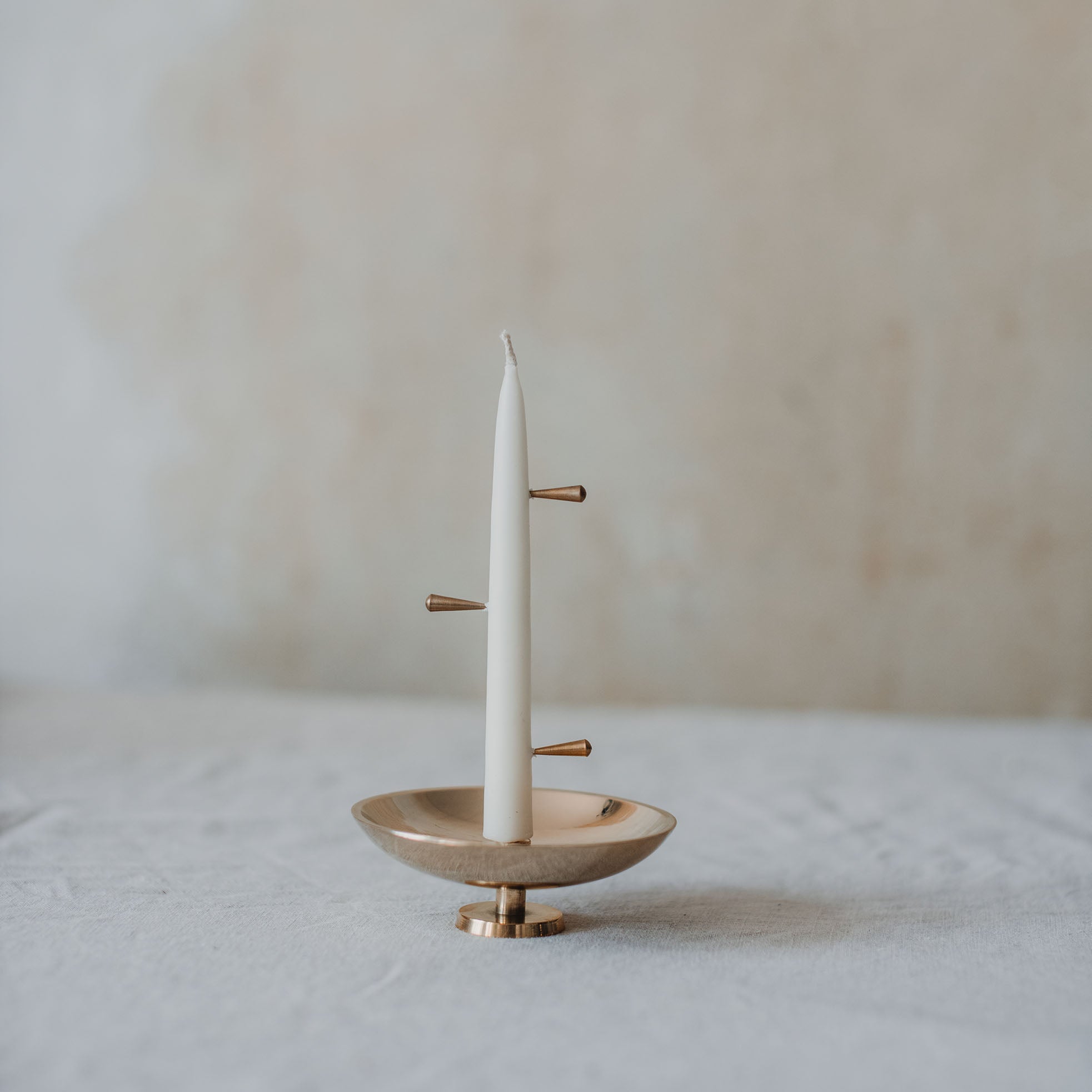 Eine weiße Stabkerze, stehend auf einer glänzenden Messingschale. In der Kerze stecken auf unterschiedlichen Höhen und Seiten kleine Messing Stifte.