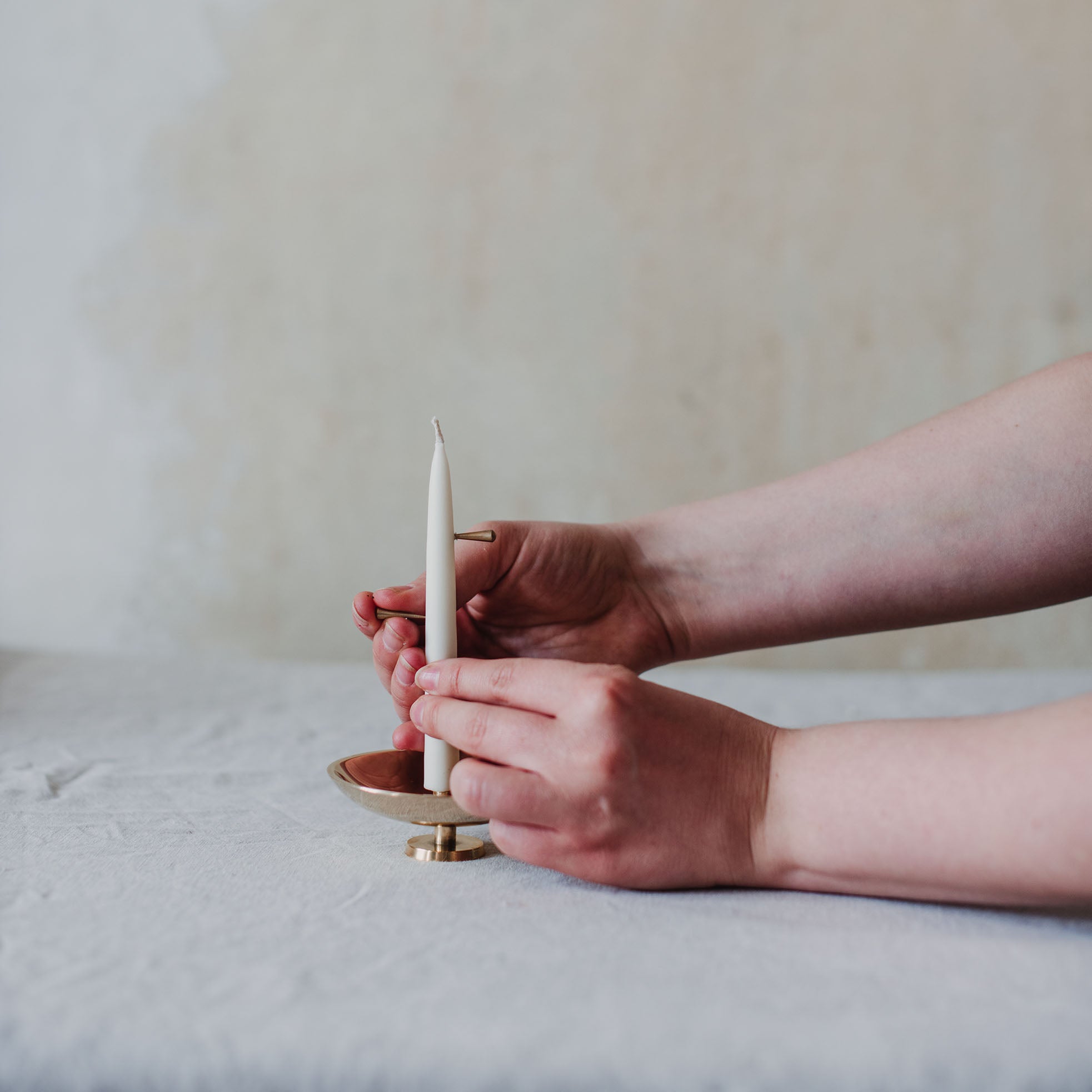 Eine weiße Stabkerze, stehend auf einer glänzenden Messingschale. Eine Person steckt einen zweiten Messingstift in die Mitte der Kerze. 