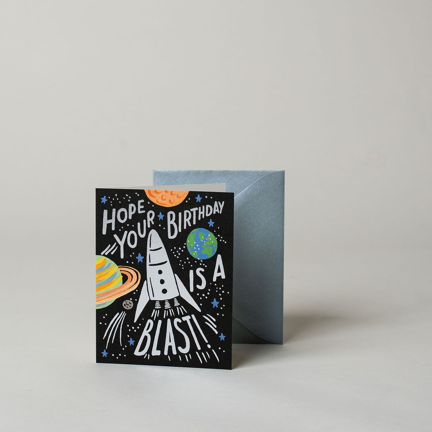 Schwarze Klappkarte mit der Aufschrift "Hope your Birthday is a blast!". In der Mitte auf der Karte ist eine Rakete abgebildet. Drumherum sind bunte Planeten illustriert.