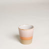 Ein Becher steht im weißen Hintergrund. Die Tasse im 70er retro Stil ist von unten nach oben pastelle grün, rosa gefärbt. In der Mitte besfindet sich ein orangener Streifen.