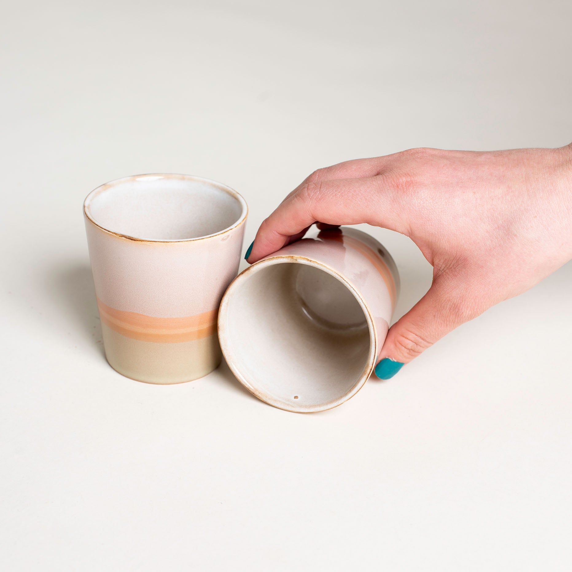 Zwei Becher in einem weißen Hintergrund, die eine stehend, die andere liegend. Die Tassen im 70er retro Stil sind von innen weiß und von außen in pastelle Fraben rosa, orange und grün gefärbt.